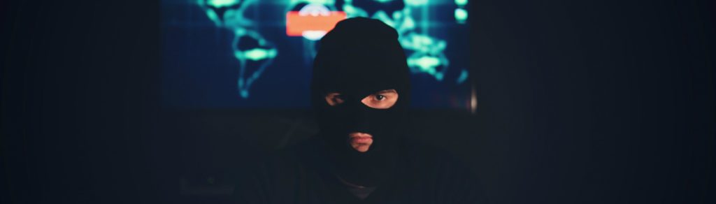 Dark photo of a person in a black balaclava looking into the camera, in front of a screen showing a glowing world map.